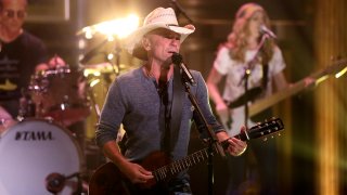 Kenny Chesney Performing on The Tonight Show with a guitar in his hand. A guitarist and drummer are behind him.