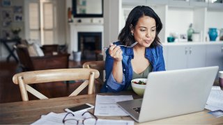 young woman eating while working from home on laptop