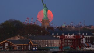 An almost full Pink Moon is seen in the sky behind the Statue of Liberty as the sun rises on April 26, 2021, in New York City. The Super Pink Moon will rise later in the day.