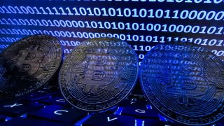 An illustration showing physical bitcoins alongside binary code displayed on a laptop.