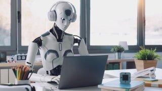 A humanoid robot works in an office on a laptop to listening music in headphones.
