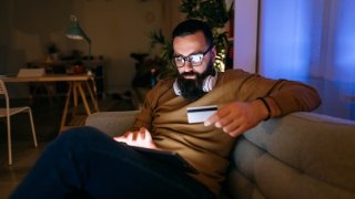 Man sits on a sofa in his living room and uses a credit card to pay online.