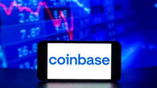 The Coinbase logo is displayed on a mobile phone screen with stock market percentages in the background.