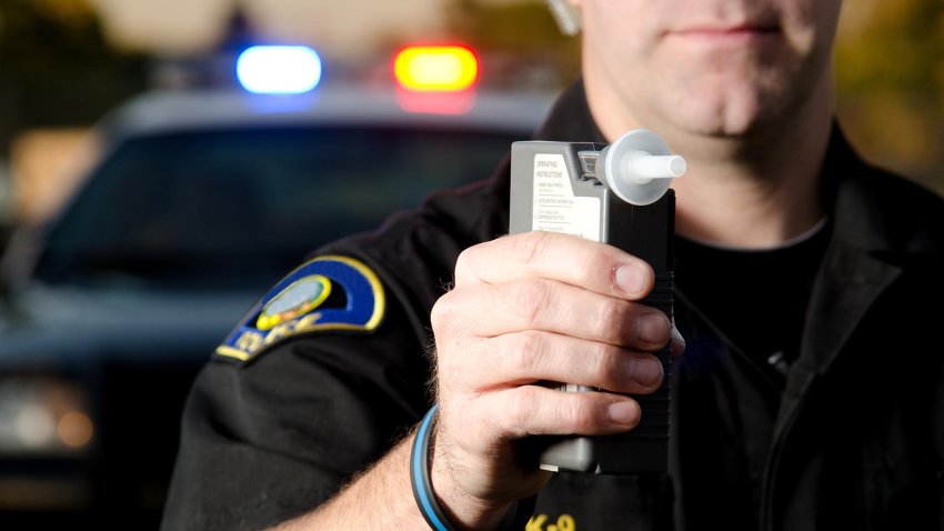 A police officer holds a breath test machine in his hand ready at a traffic stop with his patrol car in the background.