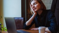 A tired young woman in a shirt sitting in a cafe, rubbing her eyes after a long day working on her laptop. A disposable coffee cup and phone lay on the table. Daylight from the window.