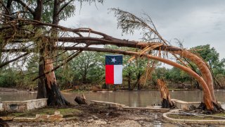 CENTER POINT, TEXAS – JULY 13: A Texas flag hangs from a storm-damaged tree on the banks of the Guadalupe River on July 13, 2025 in Center Point, Texas. More than 160 people are still missing after storm cells halted over the area, dumping nearly 15 inches of rain and causing a 22-foot rise along the Guadalupe River. (Photo by Brandon Bell/Getty Images)