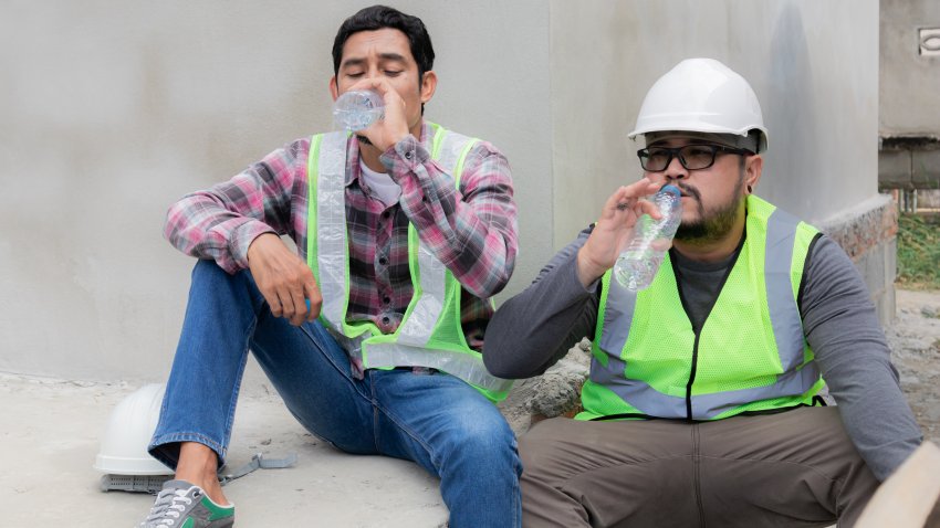 Two Asian builder workers wearing safety reflected vest and helmet tired thirsty hydrated drinking water from plastic bottle together relaxed sitting on construction site on overheated sunny day